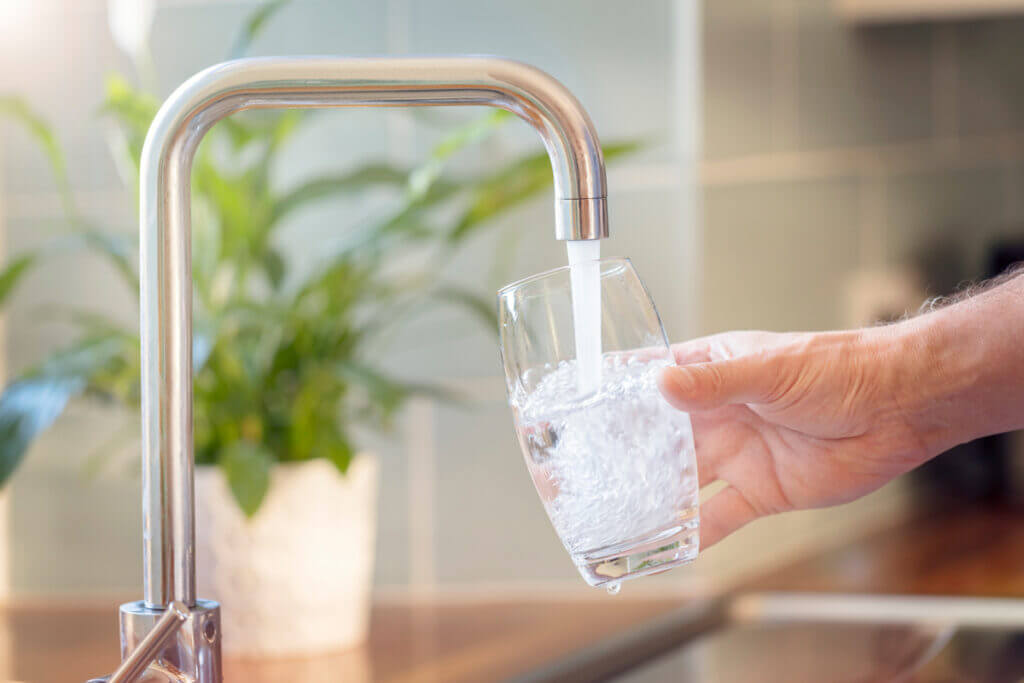 Nahaufnahme eines Wasserhahns, aus dem Trinkwasser in ein Glas fließt, das von einer Hand gehalten wird. Im Hintergrund ist eine grüne Pflanze in einem weißen Topf zu sehen.